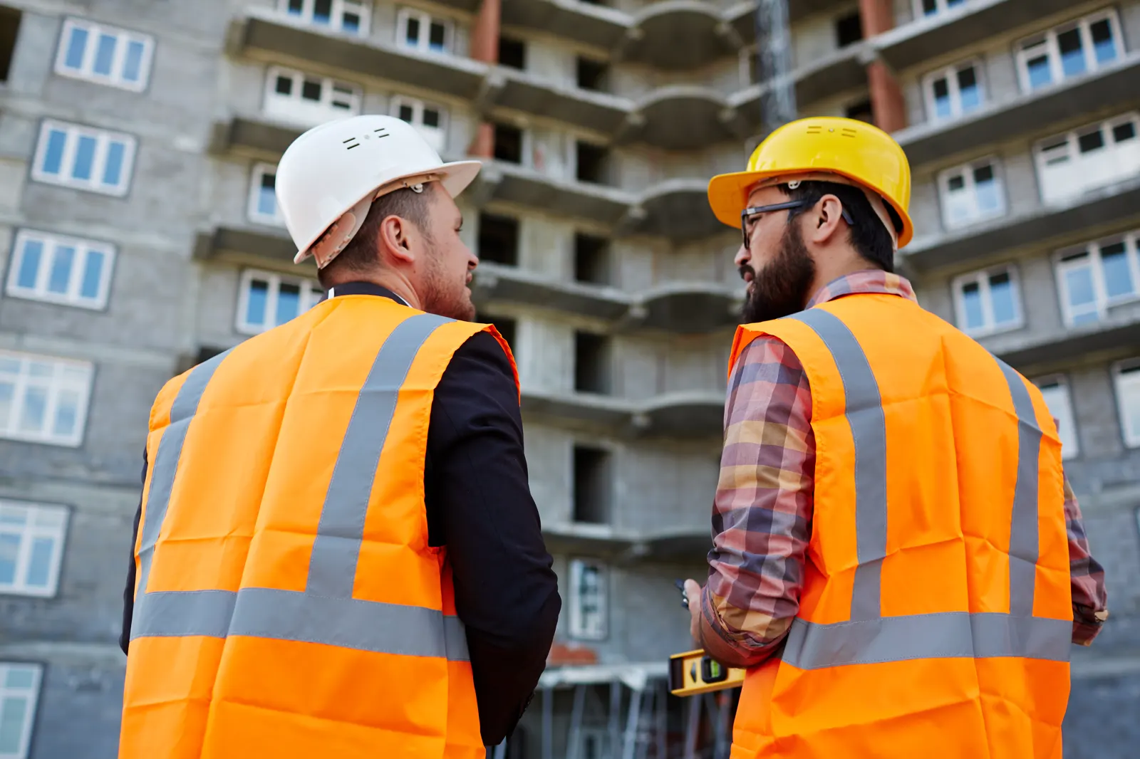 Abordagem comercial diante de uma obra em fase de estrutura dois profissionais de capacete e colete conversando diante de um prédio em construção